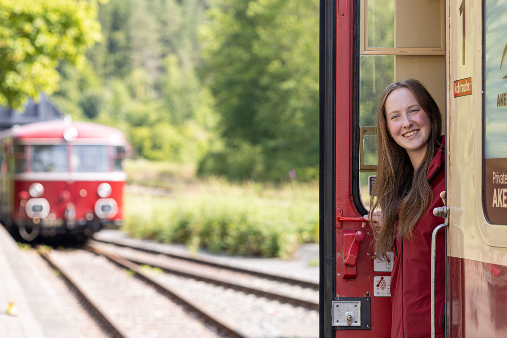 Frau lehnt aus der Tür eines roten historischen Zugwagens, im Hintergrund nähert sich ein weiterer roter Zug auf den Gleisen