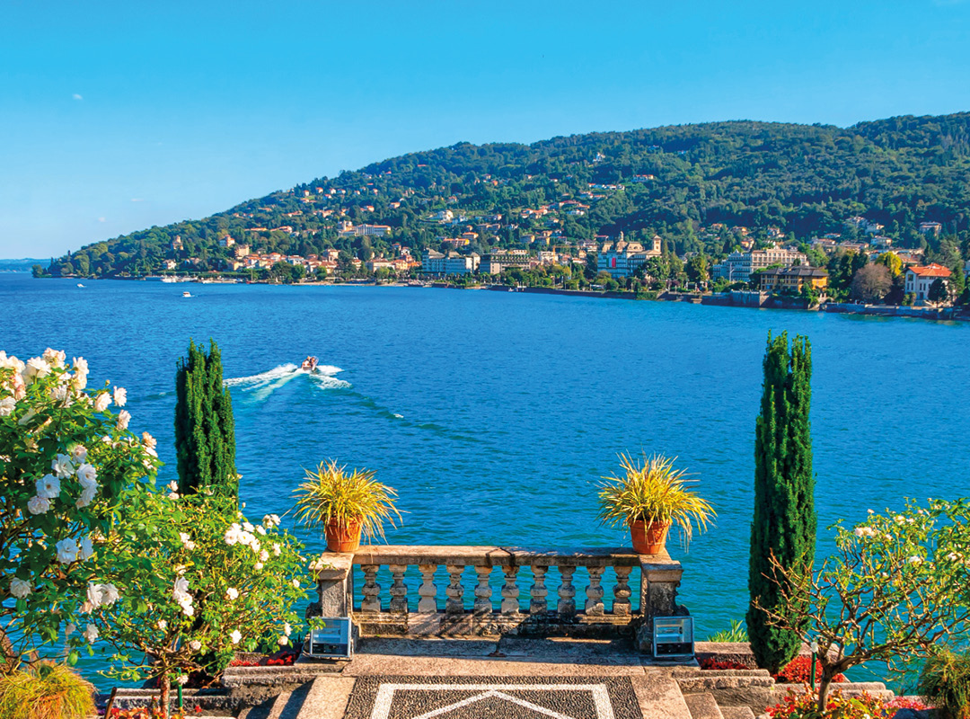 Blick von den Borromäischen Inseln auf den Lago Maggiore mit mediterraner Gartenanlage, blauem See und bewaldeter Uferlandschaft.